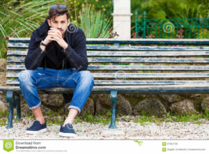 Boyfriend waiting. Handsome young man model sitting on the bench A handsome young man is sitting on a bench watching and waiting. The boy wearing trendy clothes. Beside him the empty bench. Waiting for someone or simply rest. Location: Rome, Italy. Park in the city center.