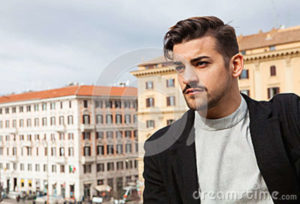 City handsome man, fashion modern hair A handsome boy looking, stylish hair and modern clothes. Horizontal portrait behind him two historic buildings in Rome, Italy. Urban scene.