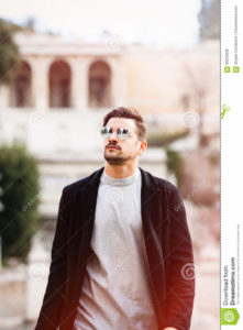 Cool handsome fashion young man. Stylish man with sunglasses A young and handsome man with sunglasses is walking in the city looking up. Trendy and modern clothing concept. The beautiful boy has a slight beard, gray shirt and black jacket. Behind him, the ancient city of Rome with blurred construction. Italy.
