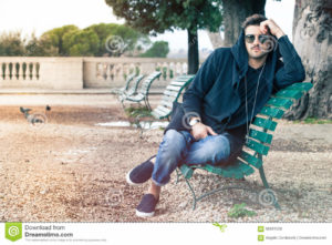 Fashionable cool young man with sunglasses relaxing on a bench A young handsome man sitting on a bench in the historic center of Rome, Italy. The boy dresses fashionable, wearing sunglasses and hoodie. Outdoors in a park.