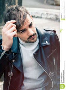 Hairstyle young man outdoors. Hair male A young and handsome guy is outdoors. He is touching the straight brown hair. Charming and youthful look. Leather jacket and light beard.