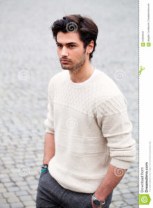 Handsome fashion young man outdoors. Black hair style. Beautiful and attractive young man with stylish hair. He is walking on the street with his hands in his pockets. White sweater and pants.