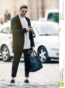 Handsome man model with sunglasses and cell phone outdoors A young and handsome man model is with mobile phone in hand, standing outdoors with urban traffic behind. The boy dresses fashionable, wearing sunglasses, trousers with a flap, a black bag in his hand. In the historic center of Rome, Italy.