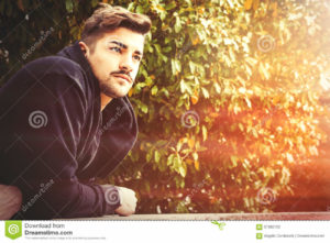 Handsome young harmony italian man - Romantic waiting A handsome young man leaning on a wall waiting for in a park with trees and leaves in the background. The young boy has a slight beard. He is wearing a coat.