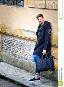 Handsome young man model on the go. Street wall Handsome young man model on the go. On the street leaning against the wall. A beautiful model with a black bag is waiting leaning against an ancient wall of an old building.