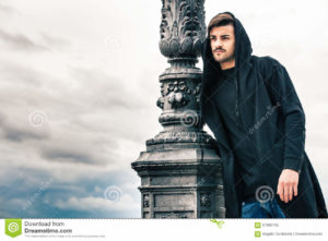 Mysterious and handsome young man model with hoody. Cloudy sky A glamorous young man is leaning against a lamppost standing outdoors. The boy is wearing a dark sweatshirt with a hood on his head. The pole is made of iron and has numerous decorations. Behind him the sky covered by clouds. Location: Rome, Italy