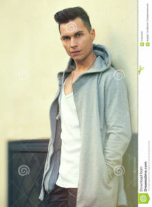 Portrait of serious young man leaning against the wall. Fashion hair style A young man dressed in youth fashion. He is leaning against a wall and stares intently. Hands in the pockets of the long hooded sweatshirt.
