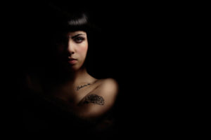 Mystery beautiful young woman in shadow and light A beautiful young girl is illuminated sideways to half by a dim light. On a black background. Bob and some tattoos on the body. The woman is naked with his arms folded. Femininity, shyness, mystery, doubts and personal care concept.