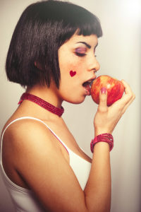 Eating a red apple. Young woman love for fruits. A young girl about to eat a red apple. The girl has a heart sticker on the cheek. Freckles and blacks bobbed hair. On clear graduated background. Love the fresh fruit, vegan diet and healthy concept.