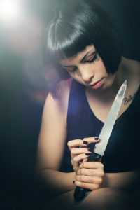 Murderous suicide girl with knife. Crime concept A beautiful girl with a knife in her hand has just committed a crime. She is touching the blade watching the knife. On the knife is reflected her look with open eyes. Black background