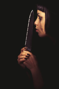 The profile of a young woman with a knife in her hand. In the darkness her face is illuminated. Crying. Home violence.