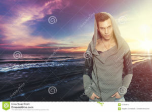 Stylish man with hooded sweatshirt the sea. Colorful sunset Stylish man with hooded sweatshirt the sea. Colorful sunset. A young man with attitude between grumpy and confidant. fashionable clothes, hooded sweatshirt. Beautiful landscape of the sea with sunset behind him.
