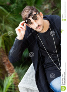 Sunglasses. Young gorgeous young man. A young handsome man discovering his eyes moving up sunglasses. Dresses the youth fashion. Chain around his neck. Outdoors.
