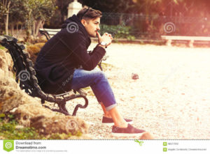 Thinking, thoughtful handsome man on the bench. Outdoors Young and handsome Italian man outdoors in a natural park in Rome, Italy. The boy is thoughtful and is thinking relaxed. Modern contemporary clothing. Trousers with turn-ups. Light warm sunbeam