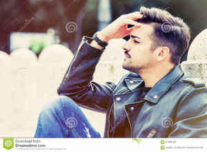 Worried young man, concern concept. Outdoor A young, beautiful and modern man is sitting with his forehead. He is thoughtful and slightly worried. Outdoors in a park, the young man has a stylish hair cut.