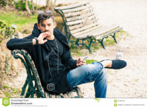 Young man sitting on the bench thinking waiting with phone in hand A handsome young boy sitting outdoors on a bench in a nature park in the city center. The young man wearing fashionable clothes youth. Waiting concept, doubts, misgivings, falling in love, social. Location: Rome, Italy.
