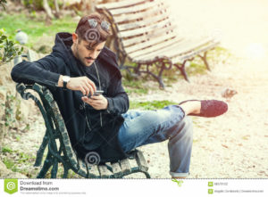 Young man sitting on the bench waiting with phone in hand A handsome young boy sitting outdoors on a bench in a nature park in the city center. The young man wearing fashionable clothes youth. Waiting concept, doubts, misgivings, falling in love, social. Location: Rome, Italy.