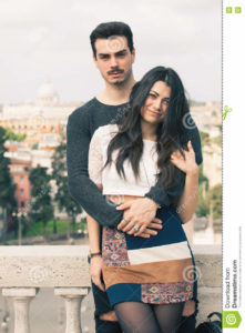 Beautiful embracing lovely young italian couple outdoors A couple, young men and a women outdoors, is embracing on a terrace in the historic center of Rome, Italy. Black hair. Bright daylight. The men has a mustache. Fashionable youth clothes.