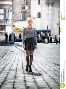 Blonde girl walking on the street in the city wearing a skirt. Girl walking on the street in the city. Beautiful young girl walking in the historic center of Rome, Italy. Driveway with paving of cobblestones. Wearing a skirt