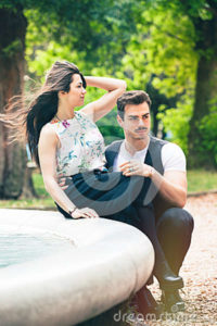Couple outdoors romance lovers in a park. Loving romantic relationship. A beautiful young couple is relaxing in a park with trees and vegetation. The girl is sitting on a fountain. The young men crouched in front of her. Hair in the wind.
