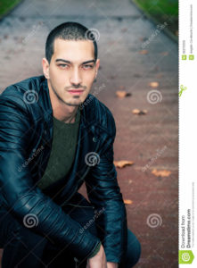 Handsome man short hair style outdoors Handsome young model outdoors. Intense look, leather jacket. Light eyes. Road with autumn scene behind him. Some leaves on the ground. Short hair style.