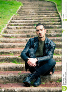 Handsome young man sitting on steps outdoors. Handsome young boy sitting on a staircase in a natural park. Crouch, rock style clothes with leather jacket and dark jeans. Short hair and earring.