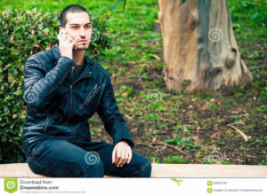 Happy young man with smartphone. Talking on the phone. Telephone conversation. Happy man outdoor. A young and handsome boy is talking on the phone with someone. His mood is serious. Outdoors in a park.