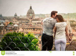 The king, his queen. Romantic couple in Rome, Italy. Two young people, a men and a women are kissing and loving. On a terrace with railing overlooking Rome, the Italian capital in Italy. The two beautiful young wearing a gray T-shirt with the words The King and His Queen. Monuments and churches with a dome on romantic background.