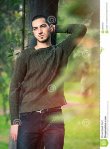 Model man short hair leaning against a tree in a nature scene A young and handsome guy leaning against a tree in nature relaxing in a natural park. Autumn fashion.