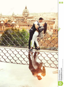 Romantic couple in Rome city, Italy. Loving relationship. Passion and love A beautiful pair embrace on a terrace with a reflection in a puddle. Behind them the historical and ancient city of Rome, Italy. Love, passionate affair, youth and genuine love concept.