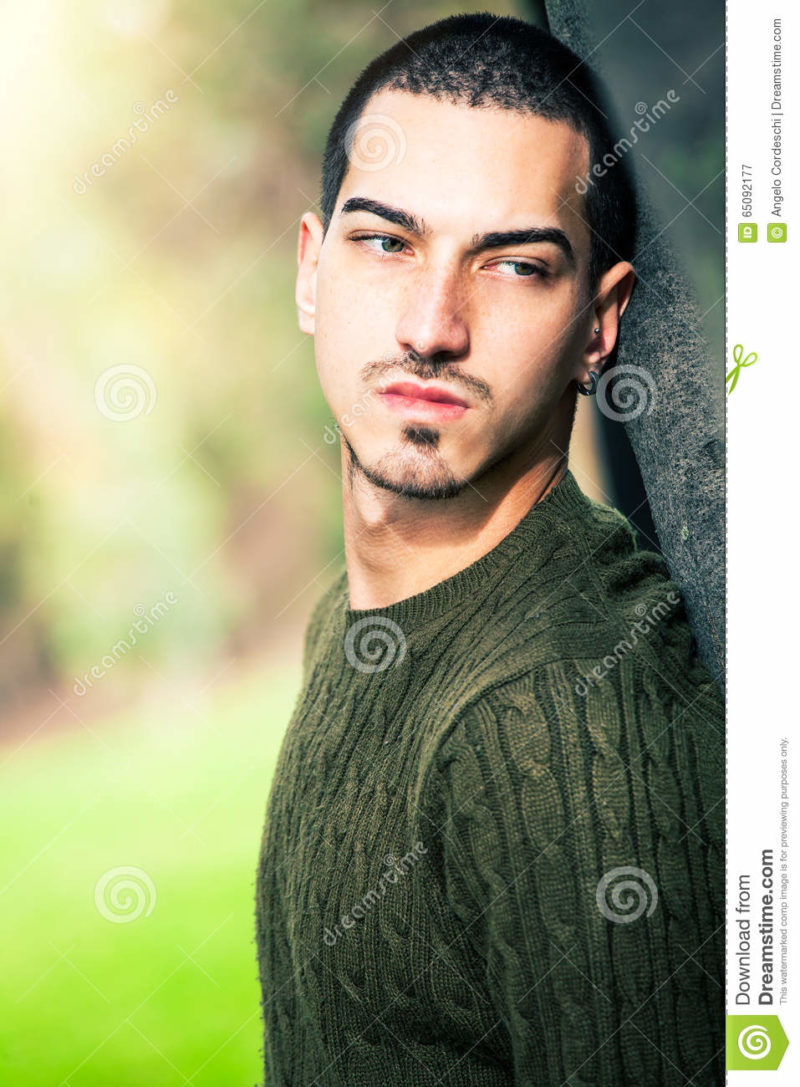 Sweet young man outdoors. Natural harmony expression. Thinking Beautiful and young boy leaning with his back on a tree. Natural background in a park. Thoughtful and relaxed attitude. The male has cropped hair and wears a green sweater.