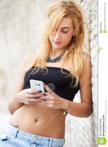 Teenage girl with mobile smartphone outdoor. Modern youth. A young blonde girl is looking at her cellphone smartphone. Modern times. Casual wear with jeans and black top. Navel piercing.