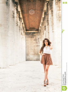 Beautiful fashion model woman curly hair with skirt A beautiful young woman is in an outdoor corridor with antique marble columns. She is fashionable. Posing. The girl is of Bolivian origin. White shirt with no sleeves and brown skirt. Lean and healthy body. Left copyspace.