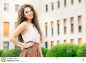 Beautiful woman with curly hair. Urban look A beautiful and happy young woman. Horizontal portrait of a girl with curly hair. Makeup on her face, perfect style. White shirt sleeveless and brown skirt. Gold bracelet. Sensuality, freshness, beauty and emotions.