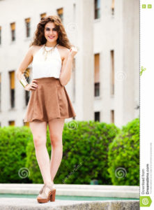 Beautiful woman with curly hair. Urban look A beautiful and happy young woman. Vertical portrait of a girl with curly hair. Makeup on her face, perfect style. White shirt sleeveless and brown skirt . Gold bracelet. Sensuality, freshness, beauty and emotions.