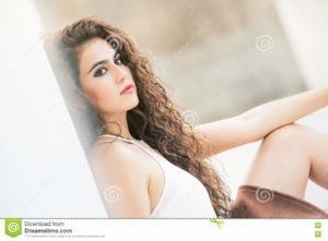Feminine beauty. Curly-haired young woman model. A young and attractive woman is leaning on a white wall. Sitting on the ground. Face with makeup. She has long curly hair. Sensual and radiant beauty. Intense emotions.