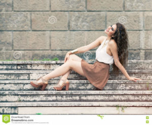 Happiness and beauty. Smiling young woman sitting on steps A young and beautiful woman smiling and joyful. She is sitting on a staircase in the historic center of Rome, Italy. Long curly hair. The girl wears a brown skirt. Wall with big bricks in the background.