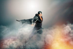 Male rock singer with cape in mysterious scenery with smoke. A black man is on a stage with smoke. Scenery with smoke and lights. Dressed in black leather.