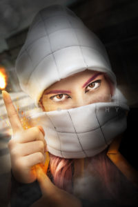 A young woman with her face covered showing only the eyes. Her hands give off energy and produce heat and flames. Cosplayer dressed as Japanese manga character.