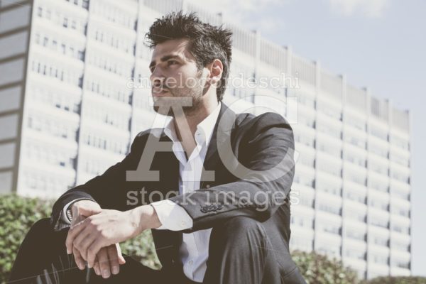 Confident handsome man sitting outdoor. Modern building behind. - Angelo Cordeschi
