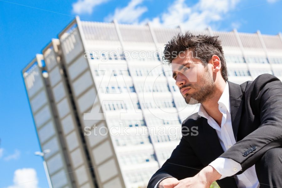 Confident handsome man sitting outdoor. Modern building behind. - Angelo Cordeschi