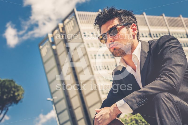 Confident handsome man with sunglasses sitting outdoor. Building behind him. - Angelo Cordeschi