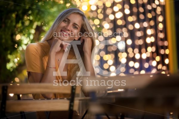 Smiling Blonde Young Woman Sitting, With Evening Fairy Lights On Angelo Cordeschi